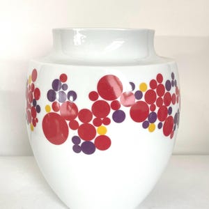 May include: White ceramic vase with a wide opening and a rounded body. The vase is decorated with a pattern of red, purple, and yellow circles. The circles are clustered together in a band around the middle of the vase.