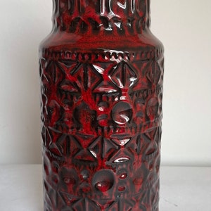 May include: A tall, cylindrical vase with a glossy, textured surface. The vase is primarily a deep red color with dark brown accents. The design features geometric and circular patterns. The vase has a wide base and a slightly narrower neck.