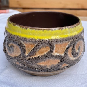 Könnte beinhalten: Eine braune Keramikschale mit gelbem Rand und einem strukturierten, abstrakten Design. Die Schale hat eine raue, fast sandige Textur.
