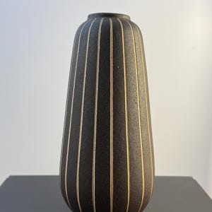 Könnte beinhalten: Eine schwarze Vase mit vertikalen, cremefarbenen Streifen. Die Vase hat eine abgerundete, längliche Form und eine schmale Öffnung. Die Streifen verlaufen über die gesamte Länge der Vase und erzeugen ein strukturiertes Aussehen. Die Vase steht auf einer dunklen Oberfläche.