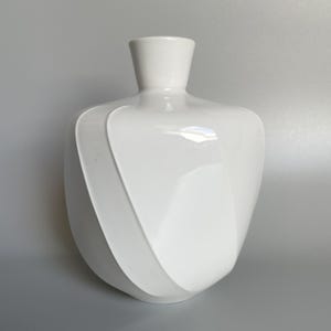 Könnte beinhalten: Eine weiße Keramikvase mit einem einzigartigen, geschwungenen Design. Die Vase hat einen breiten, abgerundeten Körper, der sich zu einem schmalen Hals verjüngt. Ein erhabenes, wirbelndes Muster umschließt die Vase. Die glänzende Oberfläche reflektiert das Licht.