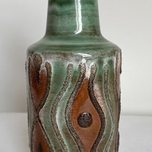 May include: A ceramic vase in shades of green and brown. The vase features a textured surface with a repeating pattern of curved lines and eye-like shapes. The base is white, and the top is a darker green.