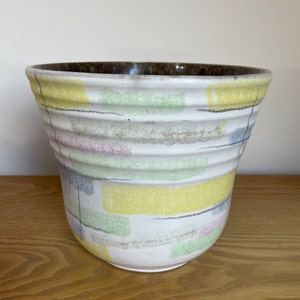 Peut inclure: Un pot en céramique blanche avec un motif géométrique dans des tons de jaune, vert, bleu et rose. Le pot a un bord brun et une lèvre légèrement évasée.