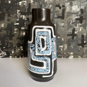 May include: A black ceramic vase with a geometric design in white and light blue. The vase has a cylindrical shape with a narrow neck and a wider base. The design features a series of interconnected squares and lines, creating a modern, abstract pattern.