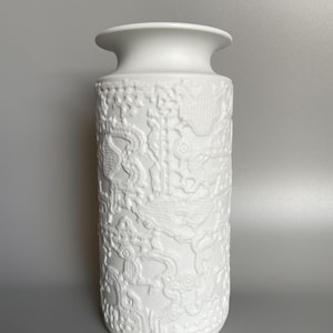 May include: White ceramic vase with an embossed floral design. The vase is tall and cylindrical.