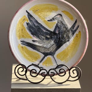 May include: A decorative ceramic plate featuring a black bird design with outstretched wings, set against a yellow and white background. The plate has a pink rim and is displayed on a decorative black metal stand.