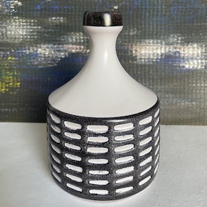 May include: A white ceramic vase with a black, textured pattern around the base. The vase has a narrow neck and a dark brown top. The background is a blurred painting with blue, green, and white colors.