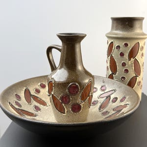 Könnte beinhalten: Ein Keramikset bestehend aus einer flachen Schale, einem kleinen Krug mit Henkel und einer hohen zylindrischen Vase. Jedes Stück hat eine gesprenkelte beige-braune Glasur mit erhabenen, glänzenden braunen Blatt- und Kreismotiven.