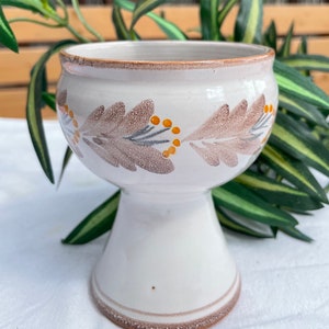 以下が含まれることがあります： 茶色と黄色の花柄が施された白い陶器の花瓶。花瓶は、幅広の丸い底と細い首をしています。