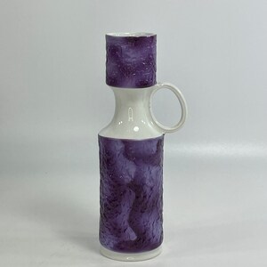May include: A white ceramic vase with a purple textured design and a handle. The vase has a wide mouth and a narrow neck.