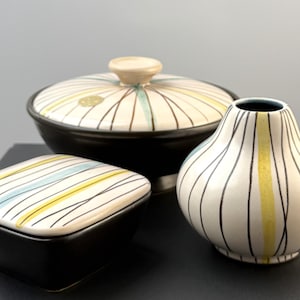 May include: A collection of three ceramic pieces including a round lidded bowl, a square lidded box, and a small vase. Each item features a cream-colored surface decorated with thin black vertical lines and accents of pale yellow and light blue.