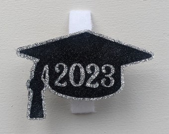 May include: Black graduation cap with silver glitter and the year 2023 in silver glitter. The cap is attached to a white clip.