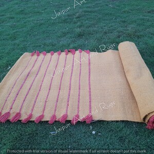 May include: A yellow and pink handwoven rug with fringe. The rug is rolled up and has a text watermark that says "Jaipur Art and Rugs".