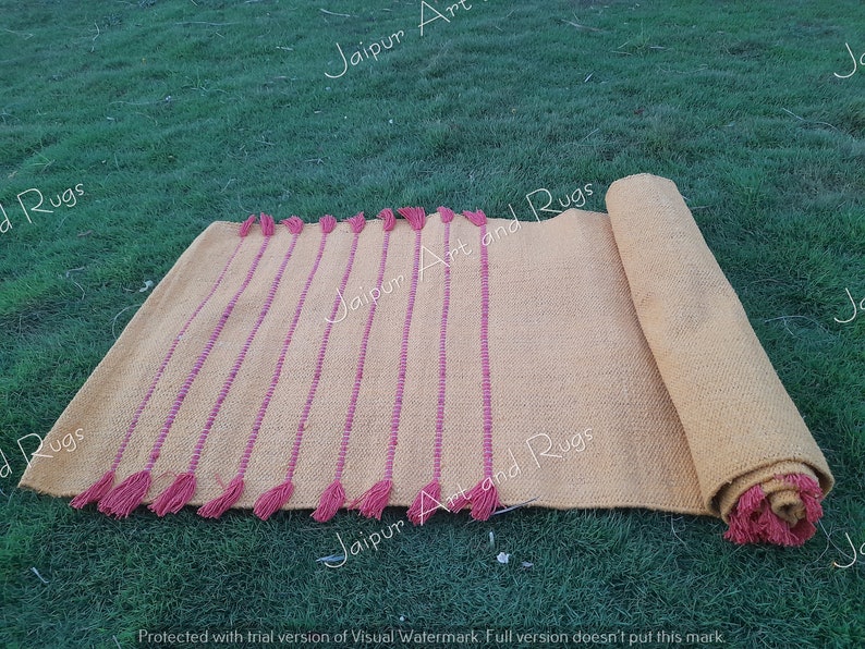 May include: A yellow and pink handwoven rug with fringe. The rug is rolled up and has a text watermark that says "Jaipur Art and Rugs".
