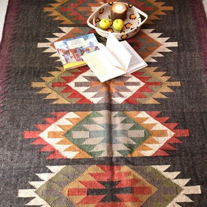 May include: A rectangular woven rug featuring a dark charcoal background with repeating geometric diamond patterns in shades of red, orange, green, and cream. The rug has fringed edges and is topped with a woven basket, two apples, and a book.
