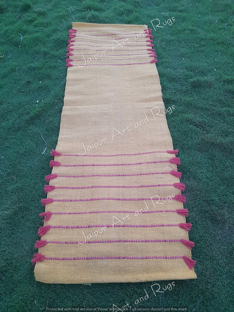 May include: A yellow and pink striped runner rug with pink tassels on each end. The rug has the text "Jaipur Art and Rugs" printed on it.