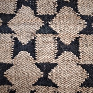 May include: A close-up of a woven rug with a geometric pattern of black and beige stars. The rug is made of natural fibers and has a textured surface.