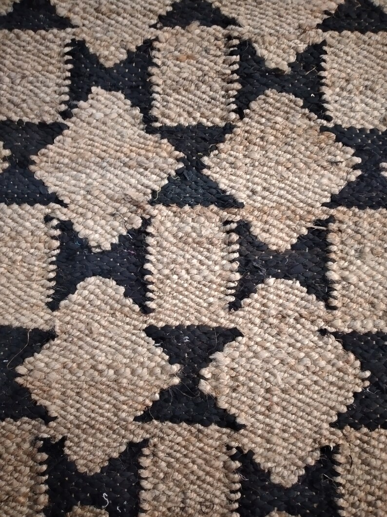 May include: A close-up of a woven rug with a geometric pattern of black and beige stars. The rug is made of natural fibers and has a textured surface.