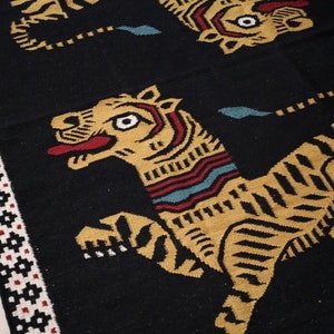 Puede incluir: Alfombra tejida en negro y dorado con un patr&oacute;n repetido de tigres estilizados. Los tigres miran hacia la derecha y tienen una lengua roja y un ojo blanco.