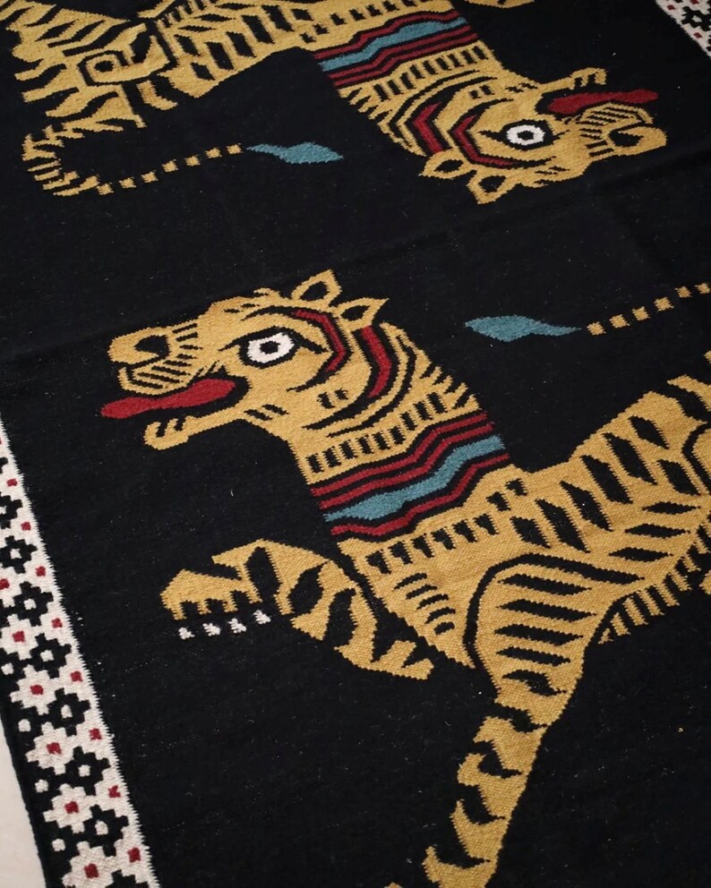 Puede incluir: Alfombra tejida en negro y dorado con un patr&oacute;n repetido de tigres estilizados. Los tigres miran hacia la derecha y tienen una lengua roja y un ojo blanco.