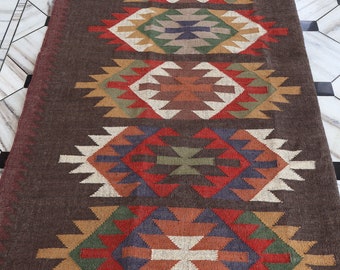 May include: A brown and multi-colored geometric patterned rug with fringe on the ends. The rug has a dark brown background with red, green, blue, and white geometric patterns.