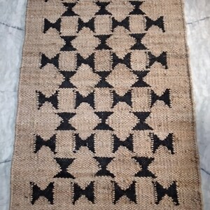 May include: A brown and black jute rug with a geometric pattern. The rug has a diamond shape with a black bow tie in the center of each diamond.
