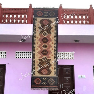 May include: A black and brown handwoven rug with a geometric pattern hangs on a wall outside a building. The rug is made of jute and wool and has a fringe on the bottom. The building is pink and has white trim. The rug is labelled "Jaipur Art and Rugs".
