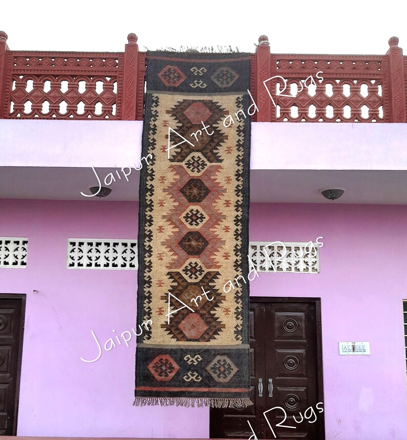 May include: A black and brown handwoven rug with a geometric pattern hangs on a wall outside a building. The rug is made of jute and wool and has a fringe on the bottom. The building is pink and has white trim. The rug is labelled "Jaipur Art and Rugs".