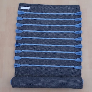Op de afbeelding: Een rechthoekig donker antracietgrijs geweven kleed met tien horizontale lichtblauwe strepen. Elke streep eindigt aan beide kanten in een klein lichtblauw kwastje. Een wit rechthoekig label met zwarte tekst is bevestigd in de linkerbovenhoek.