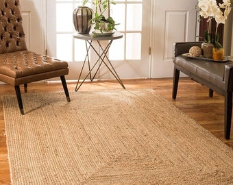 May include: A natural fiber rug with a woven texture in a light brown color. The rug has a border that is slightly darker than the center. The rug is on a wooden floor in a living room setting.