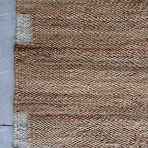 May include: Close-up of a woven jute rug with a natural brown color. The rug has a textured surface with a repeating pattern. The edge of the rug features a white border with rectangular accents.
