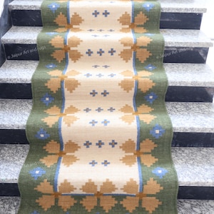 Puede incluir: Una alfombra rectangular para escaleras con un campo central crema y un borde verde. La alfombra presenta patrones geométricos en tonos marrones y azules. La alfombra está colocada sobre una escalera de mármol gris y negro.