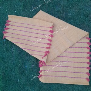 May include: Three beige handwoven placemats with pink stripes and pink tassels. The placemats are on a green grassy surface. The text "Jaipur Art and Rugs" is visible on the placemats.