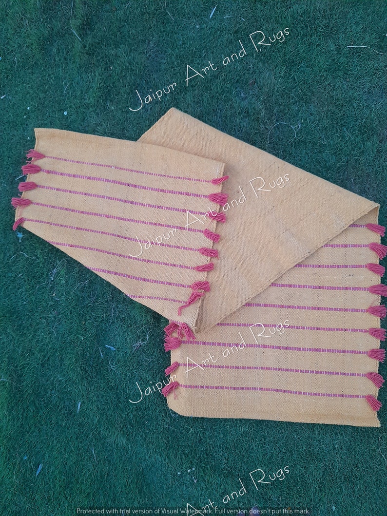 May include: Three beige handwoven placemats with pink stripes and pink tassels. The placemats are on a green grassy surface. The text "Jaipur Art and Rugs" is visible on the placemats.