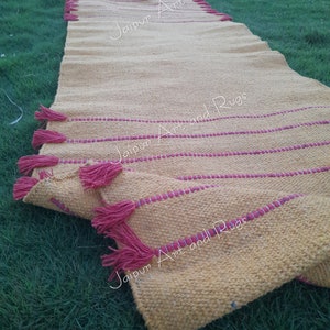 May include: A yellow handwoven rug with red fringe and red stripes. The rug is folded and lying on green grass. The text "Jaipur Art and Rugs" is visible on the rug.