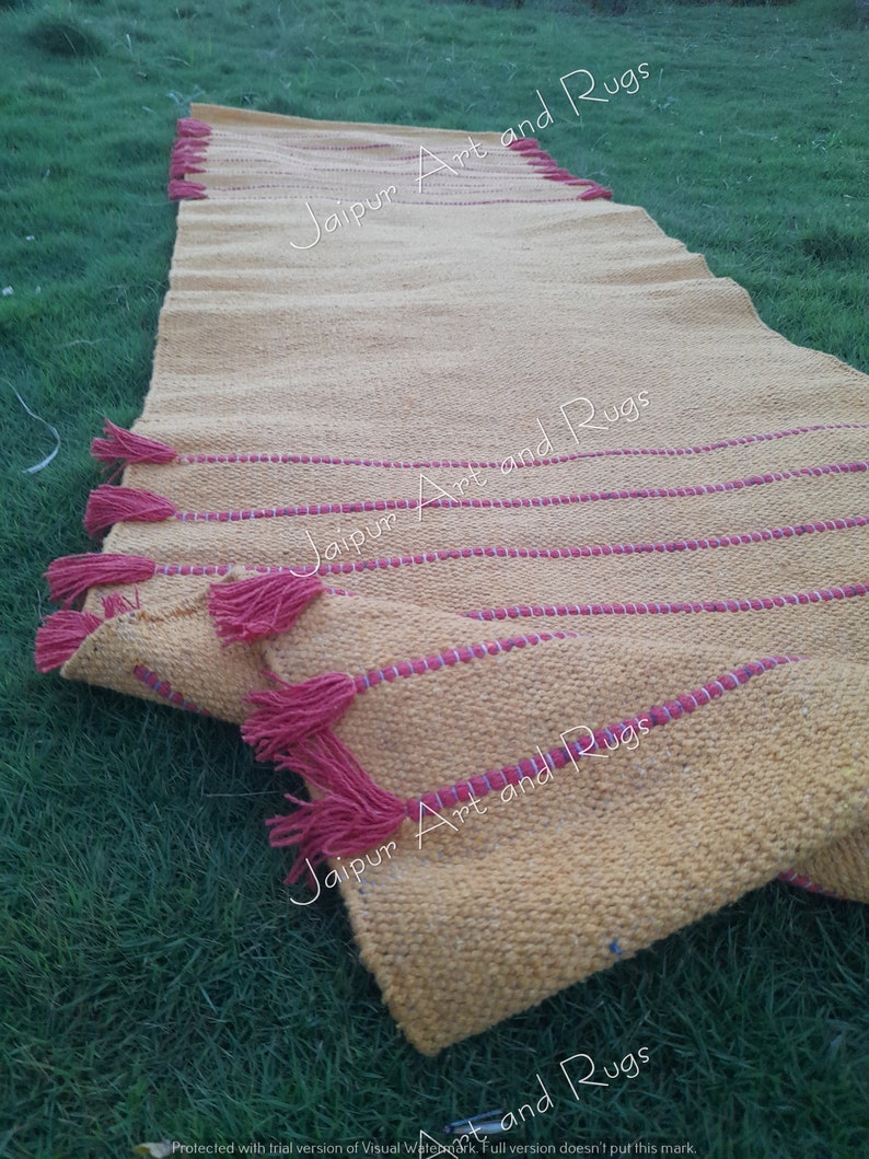 May include: A yellow handwoven rug with red fringe and red stripes. The rug is folded and lying on green grass. The text "Jaipur Art and Rugs" is visible on the rug.