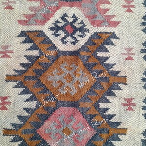 May include: A close-up of a woven rug with a geometric pattern in shades of brown, grey, pink, and white. The rug has a traditional design with repeating triangles and diamonds. The text "Jaipur Art and Rugs" is repeated in the image.