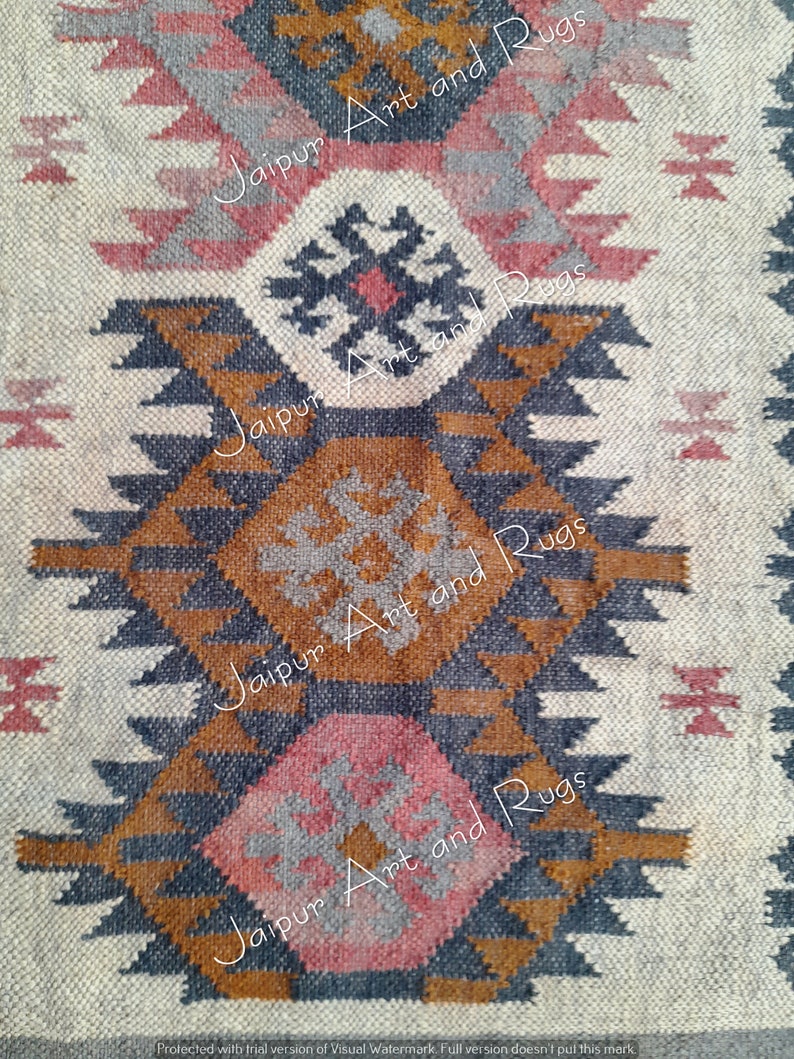May include: A close-up of a woven rug with a geometric pattern in shades of brown, grey, pink, and white. The rug has a traditional design with repeating triangles and diamonds. The text "Jaipur Art and Rugs" is repeated in the image.