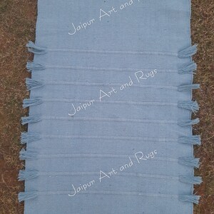 May include: A light blue cotton rug with fringe on the edges. The rug has the text "Jaipur Art and Rugs" printed on it three times.