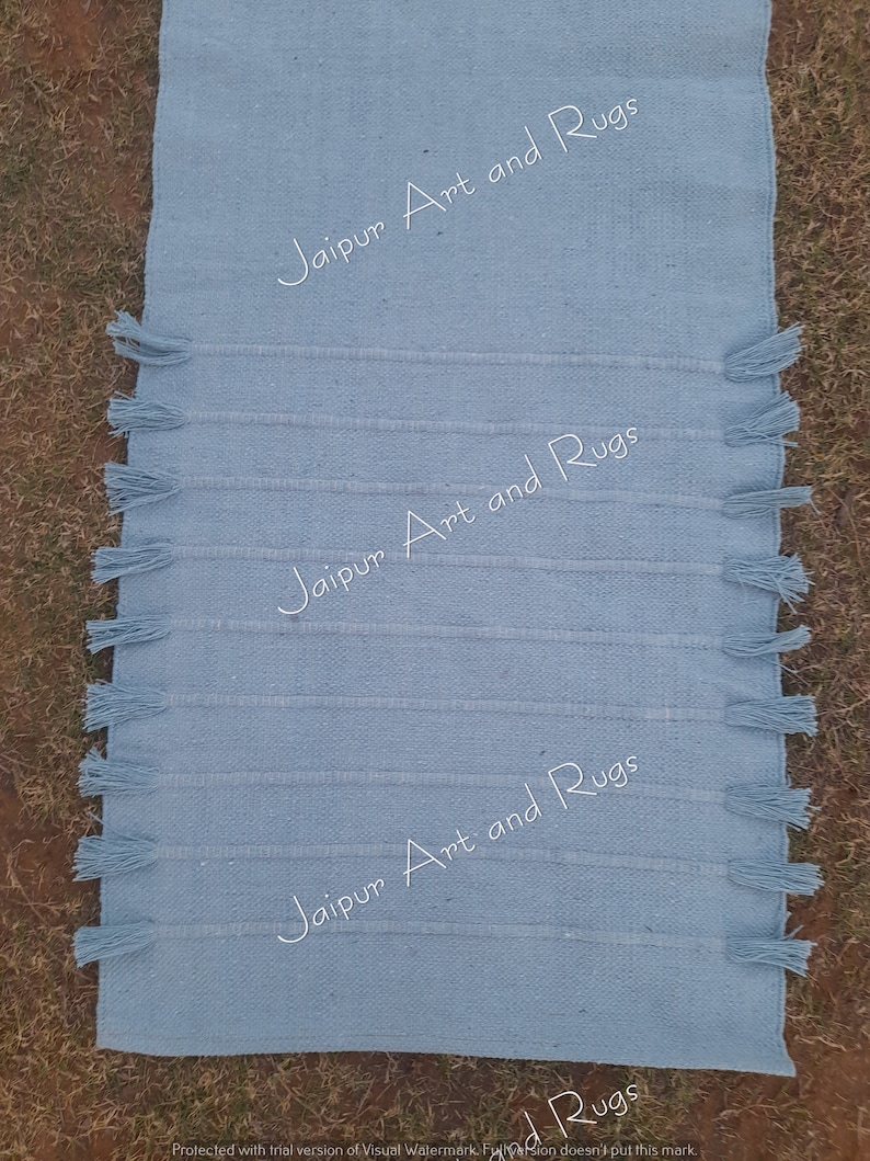 May include: A light blue cotton rug with fringe on the edges. The rug has the text "Jaipur Art and Rugs" printed on it three times.