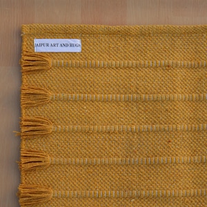 Op de afbeelding: Een mosterdgeel geweven tapijt met een reeks horizontale, getextureerde strepen en bijpassende kwastjes langs de rand. Een wit rechthoekig stoffen label met de zwarte tekst JAIPUR ART AND RUGS is in de linkerbovenhoek van het tapijt genaaid.