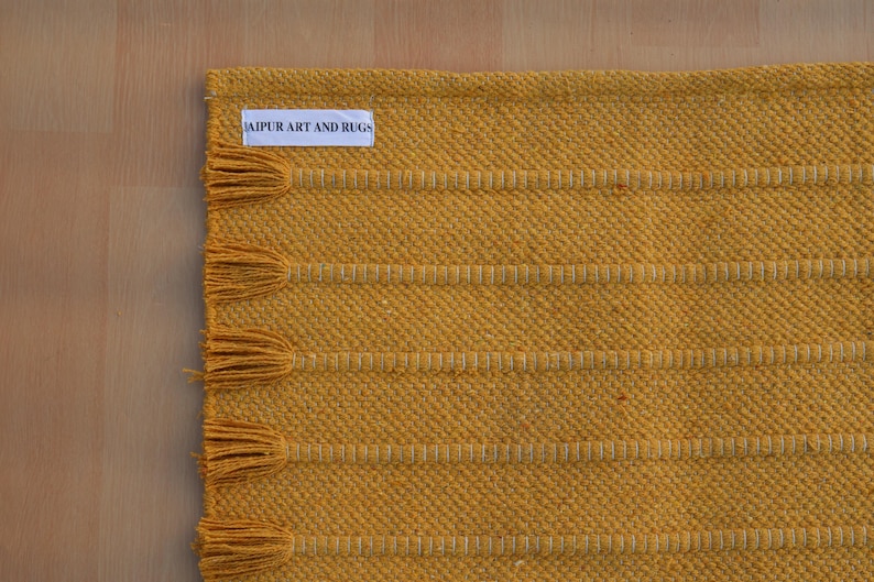 Op de afbeelding: Een mosterdgeel geweven tapijt met een reeks horizontale, getextureerde strepen en bijpassende kwastjes langs de rand. Een wit rechthoekig stoffen label met de zwarte tekst JAIPUR ART AND RUGS is in de linkerbovenhoek van het tapijt genaaid.
