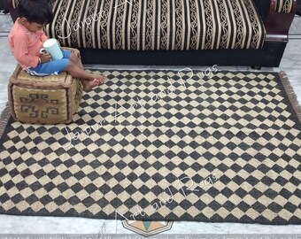 May include: A black and beige checkered rug with fringe edges. The rug is made of natural fibres and has a rustic look.