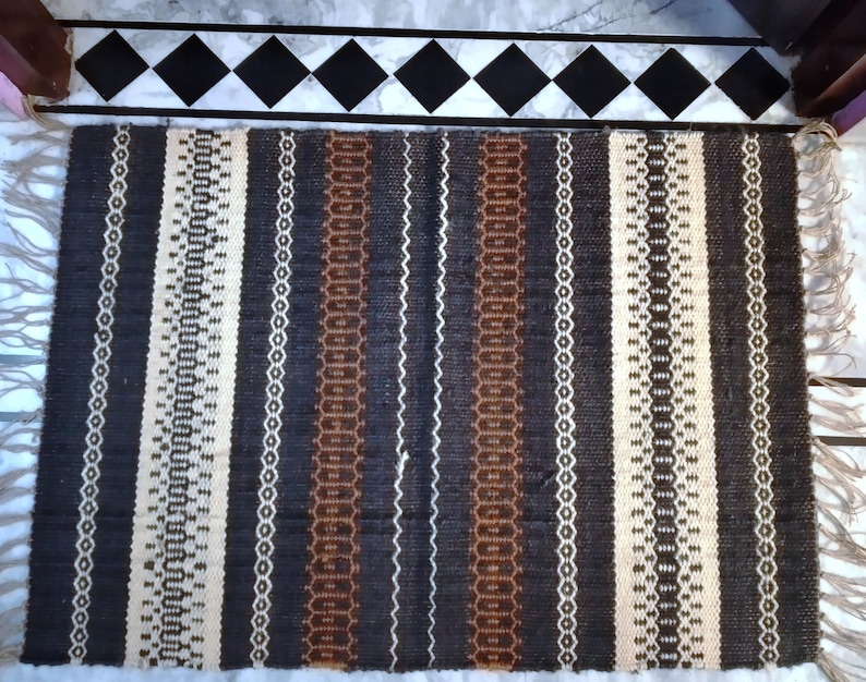 May include: A woven rug with a striped pattern in shades of brown, black, and white. The rug has a fringe on both ends.