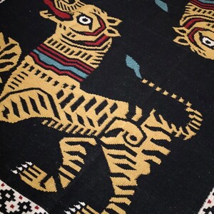Puede incluir: Alfombra negra con un patr&oacute;n repetido de tigres estilizados en dorado y marr&oacute;n. La alfombra tiene un borde negro con un patr&oacute;n geom&eacute;trico en blanco, rojo y negro.