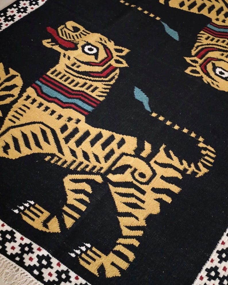 Puede incluir: Alfombra negra con un patr&oacute;n repetido de tigres estilizados en dorado y marr&oacute;n. La alfombra tiene un borde negro con un patr&oacute;n geom&eacute;trico en blanco, rojo y negro.