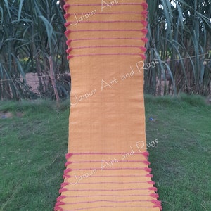 May include: A long, yellow rug with pink tassels and stripes. The rug has the text "Jaipur Art and Rugs" written on it.
