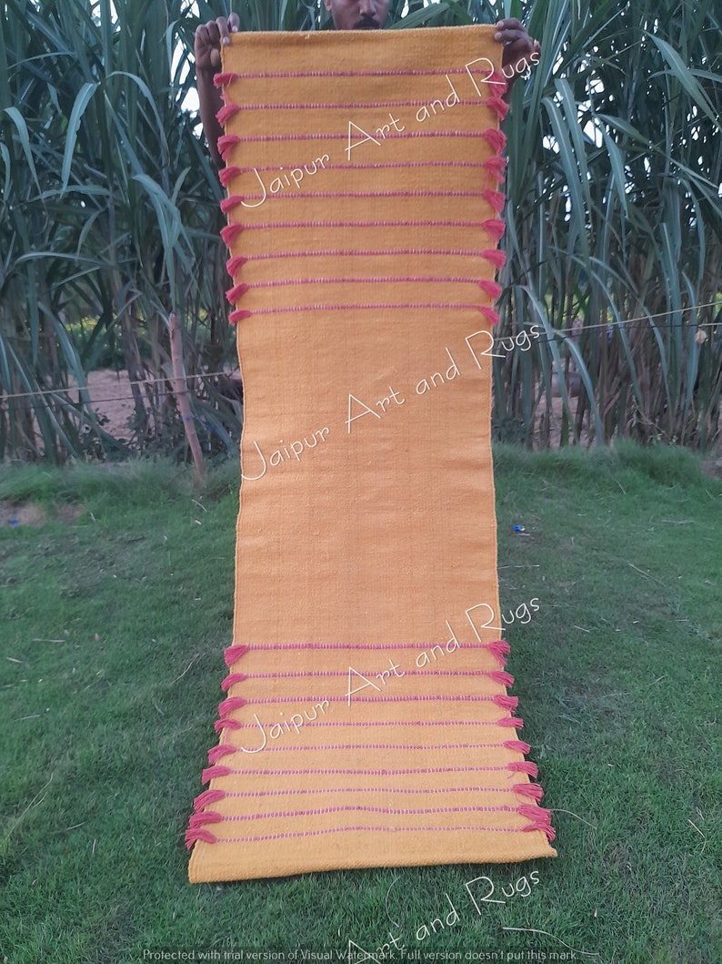 May include: A long, yellow rug with pink tassels and stripes. The rug has the text "Jaipur Art and Rugs" written on it.