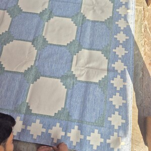 May include: A rectangular area rug featuring a repeating geometric pattern of large off-white squares separated by blue and sage green stepped borders. The rug includes a wide border with a repeating white cross motif on a blue background.