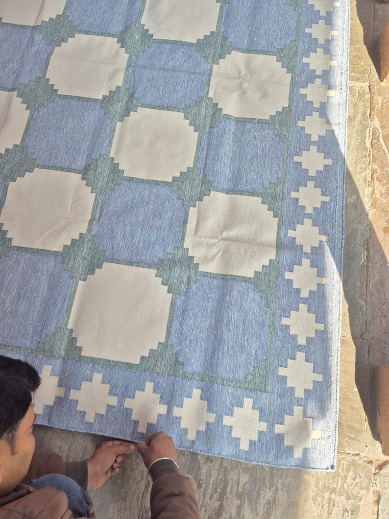 May include: A rectangular area rug featuring a repeating geometric pattern of large off-white squares separated by blue and sage green stepped borders. The rug includes a wide border with a repeating white cross motif on a blue background.